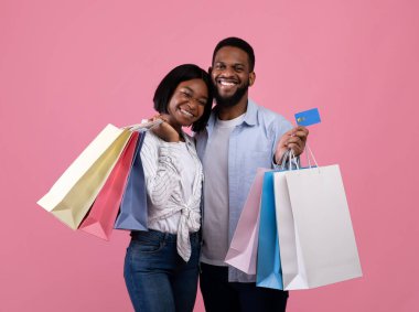 Kredi kartı ve alışveriş çantaları olan, gülümseyen ve pembe stüdyo arka planında poz veren Afro-Amerikan çift.