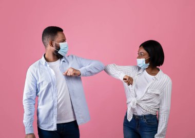 Koronavirüsün yayılmasını önlemek için yeni bir selamlama. Yüz maskeli Afro-Amerikalı çift pembe arka planda dirseklerini çarpıştırıyor.