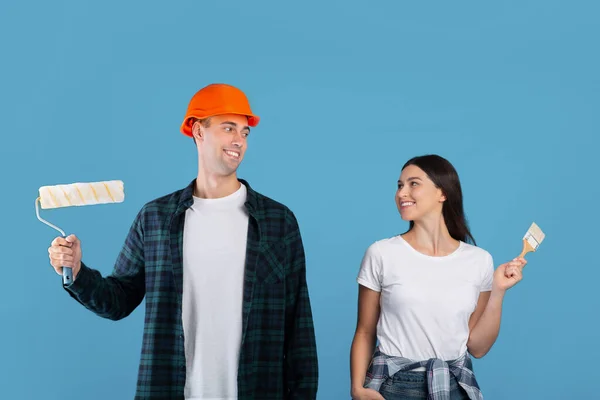 Feliz pareja joven con herramientas de pintura en las manos posando sobre fondo azul — Foto de Stock