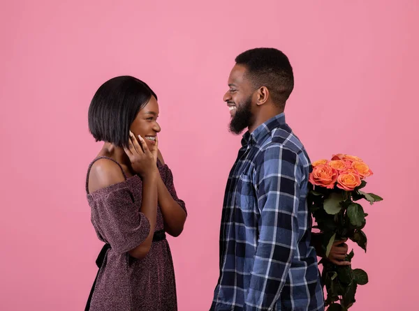 Zijaanzicht van positieve zwarte man die bloemen verbergt voor zijn geliefde achter zijn rug over roze achtergrond. Valentijnsdag — Stockfoto