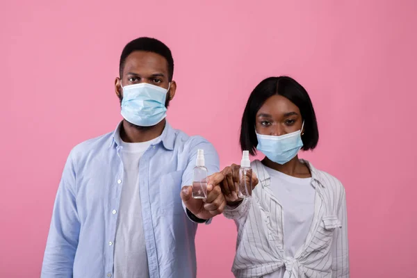 Millennial casal preto em máscaras médicas segurando garrafas de anti-séptico em fundo de estúdio rosa, foco seletivo — Fotografia de Stock