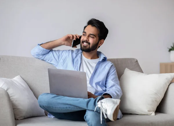 Trabajo remoto. Hombre árabe usando el ordenador portátil y hablando en el teléfono celular en casa — Foto de Stock