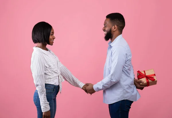 Kochający czarny facet trzymający swoją ukochaną dłoń, chowający prezent dla niej za plecami nad różowym tłem, widok z boku — Zdjęcie stockowe