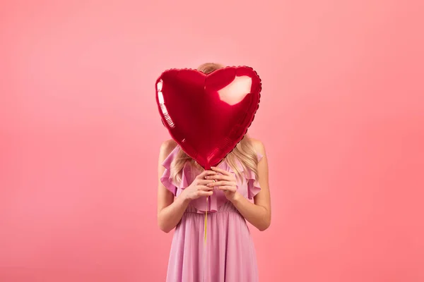 Молода жінка покриває обличчя кулею у формі серця на рожевому фоні студії. День Святого Валентина — стокове фото
