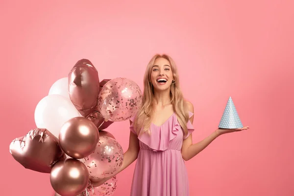 Dokonalé narozeniny. Mladá okouzlující žena v krásné šaty drží B-day klobouk a prázdninové balónky přes růžové pozadí — Stock fotografie