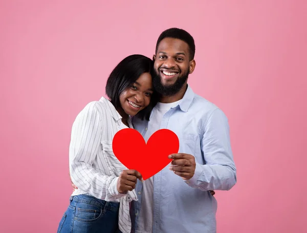Portrait of passionate black guy and his happy girlfriend hugging, holding red paper heart on pink studio background — Stock Photo, Image