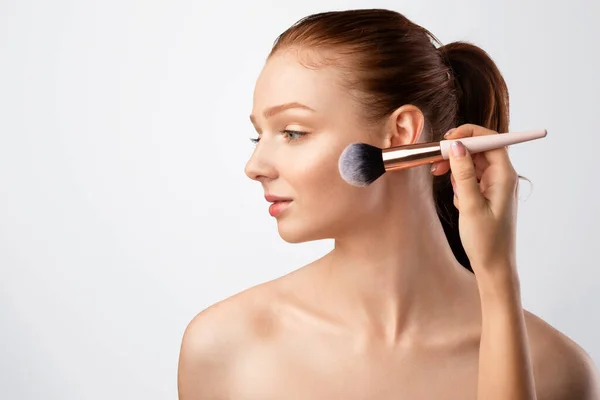 Fille aux cheveux rouges faisant maquillage avec brosse regardant à l'extérieur, fond blanc — Photo