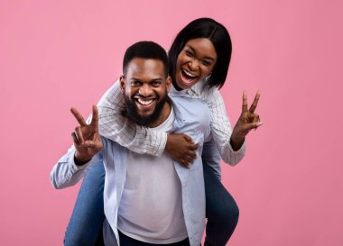 Yakışıklı siyahi adam, sevgilisini sırtında taşıyor. Pembe stüdyo geçmişinde barış ya da zafer işareti yapıyor.