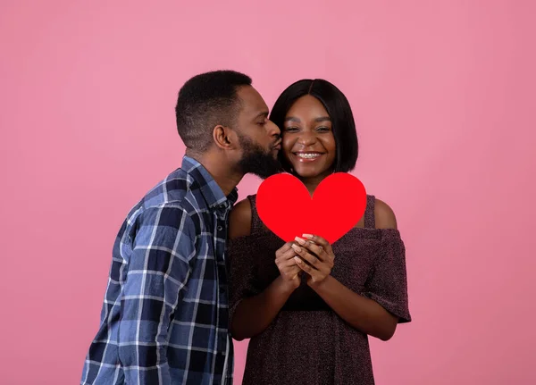 Celebração do Dia dos Namorados. Bonito cara preto beijando sua bela namorada com coração vermelho no fundo rosa — Fotografia de Stock