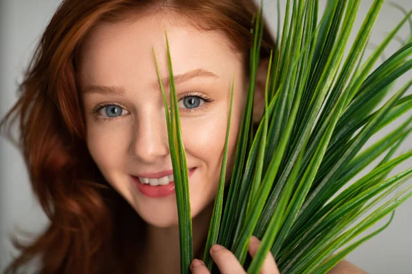 Roodharige jonge vrouw poseren met groene plant over grijze achtergrond — Stockfoto