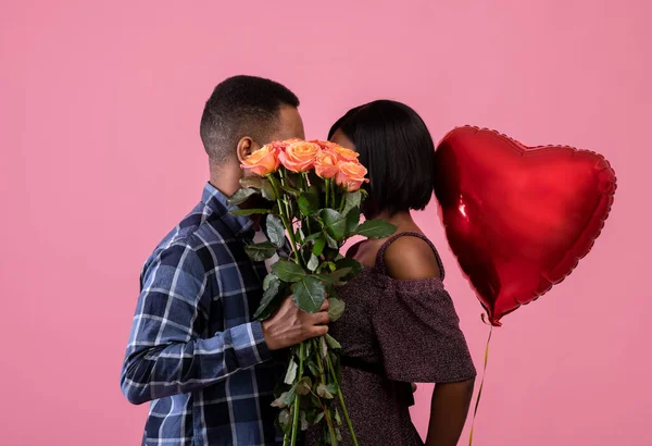Kochająca Afroamerykanka para całuje się za bukietem róż, trzymając balon w kształcie serca na różowym tle studio — Zdjęcie stockowe