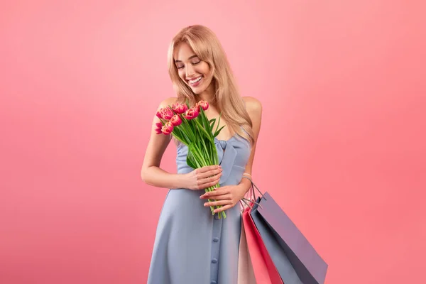 Ventes de la Journée internationale des femmes. Jolie jeune femme avec bouquet de tulipes et sacs à provisions sur fond de studio rose — Photo