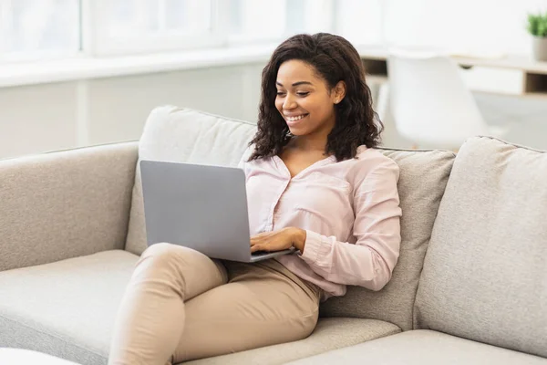 Donna nera seduta sul divano e al lavoro sul computer portatile — Foto Stock