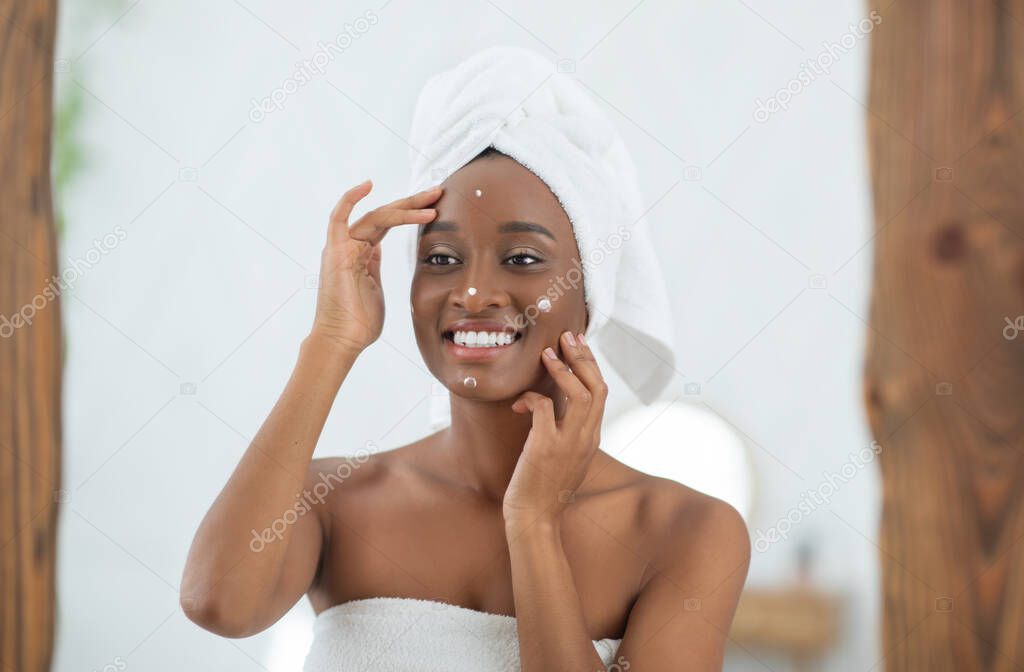 Happy african american lady with white towel on head looks in mirror and smears face with cream