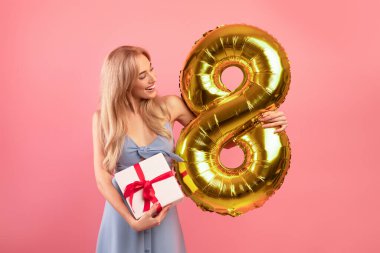Elinde sekiz numaralı altın balon ve pembe stüdyo arka planında bir buket taze bahar lalesi tutan mutlu genç bayan.