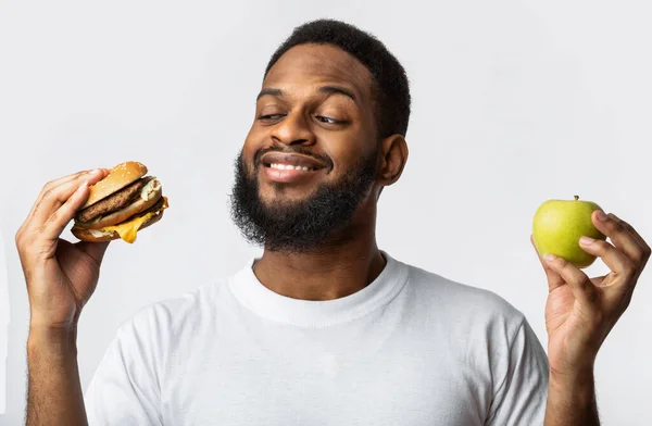 Preto cara segurando hambúrguer e maçã escolhendo comida, fundo branco — Fotografia de Stock