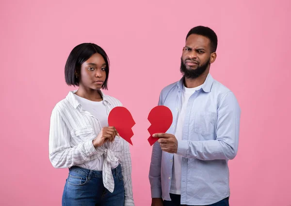 Fin del concepto de amor. Infeliz joven pareja negra sosteniendo pedazos de corazón roto en el fondo del estudio rosa —  Fotos de Stock