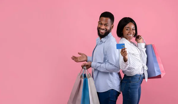 Positieve zwarte vrouw met vriendje houden geschenkzakjes en credit card, poseren en kijken naar de camera op roze achtergrond — Stockfoto