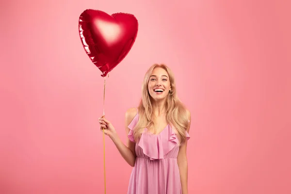 Portrét radostné blondýny v šatech drží červené srdce ve tvaru balónu na Valentýna na růžové pozadí studia — Stock fotografie