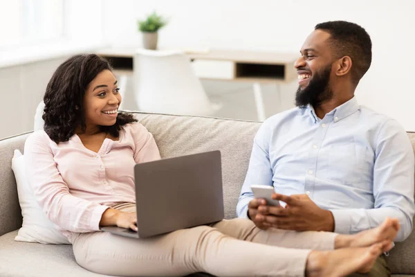 Africano casal americano sentado no sofá, usando laptop e smartphone — Fotografia de Stock