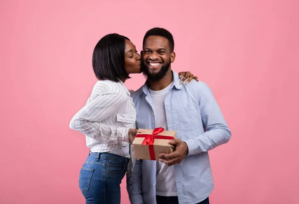 Любящая чёрная женщина дарит своему счастливому мужу Валентину подарок, целует его в щеку на фоне розовой студии — стоковое фото