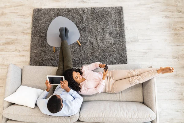 African american couple sitting on couch, using tablet and smartphone