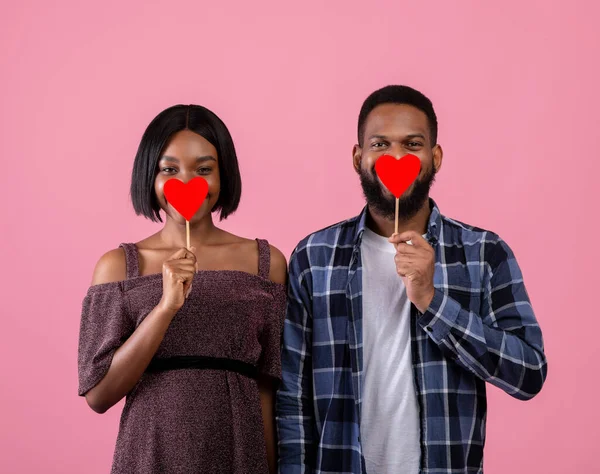 Happy Valentines Day. Passionate black couple covering their mouths with red hearts on pink studio background — Stock Photo, Image