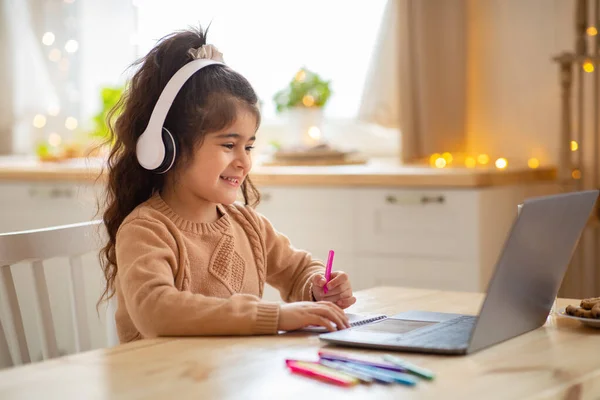 Koncepce domácího vzdělávání. Usmívající se holčička ve sluchátkách studovat doma s notebookem — Stock fotografie