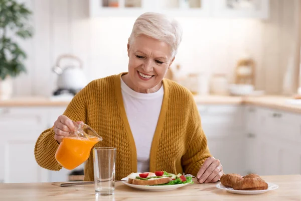 Atractiva anciana sirviendo jugo, almorzando en la cocina — Foto de Stock