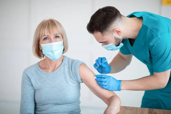 Dokter in medisch masker geeft covid-19 vaccinschot aan oudere vrouwelijke patiënt tijdens immunisatiecampagne in het ziekenhuis — Stockfoto