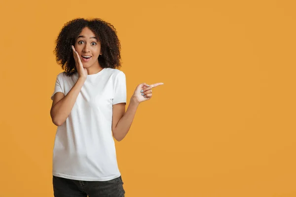 Sorprendido joven afroamericano alegre mujer en camiseta blanca presiona mano a mejilla y señala el espacio vacío — Foto de Stock