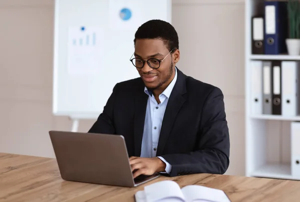 Молодой черный предприниматель, работающий с ноутбуком, офисным интерьером — стоковое фото