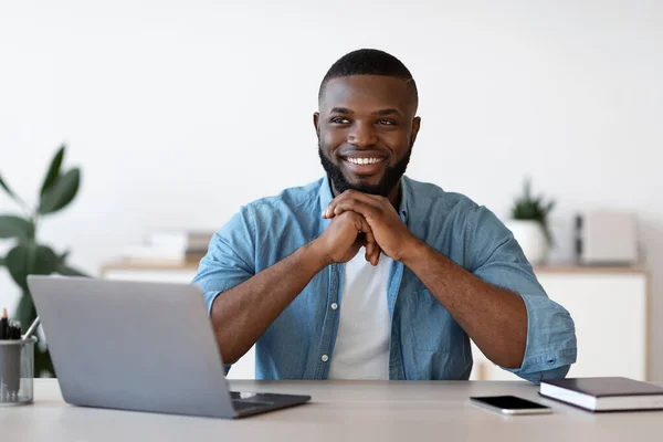 Trabalhador negro pensivo do escritório do homem que senta-se na mesa com portátil e sorrindo — Fotografia de Stock