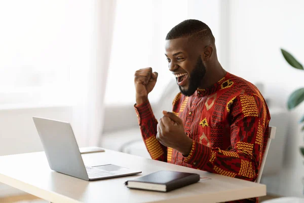 Online výhra. Radostný černoch v tradiční košili slaví úspěch s notebookem — Stock fotografie