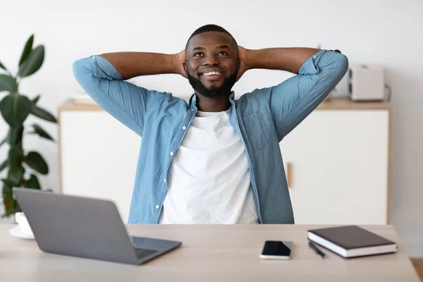 Autónomo masculino negro positivo que se inclina hacia atrás en la silla, descansando en el lugar de trabajo — Foto de Stock