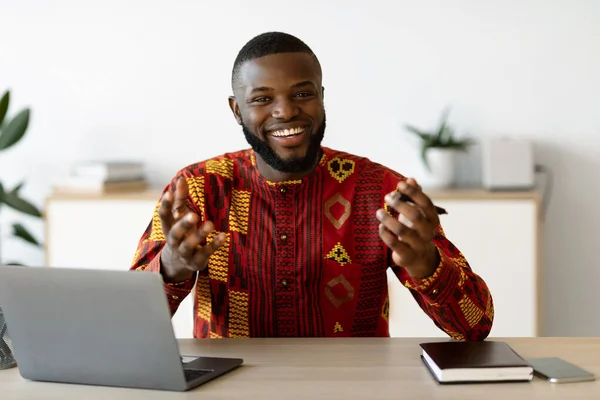 Radostný africký muž v tradičním kostýmu sedí u stolu a mluví na kameře — Stock fotografie