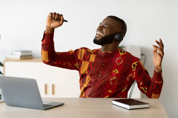 Alegre freelancer africano divirtiéndose en el lugar de trabajo, cantando y escuchando música —  Fotos de Stock