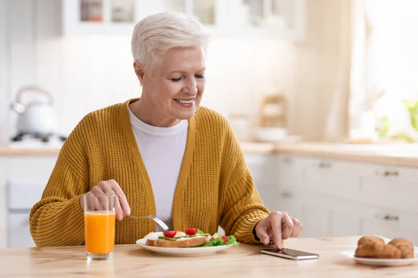 Feliz anciana usando un teléfono inteligente moderno mientras almuerza — Foto de Stock