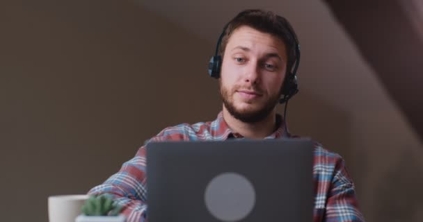 Молодой человек в гарнитуре, наблюдающий онлайн веб-бинар или живую встречу на ноутбуке, выглядящий заинтересованным и сосредоточенным — стоковое видео