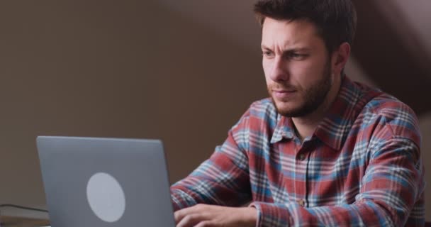 Joven hombre serio pensando en la nueva estrategia de marketing, pensando y escribiendo en el ordenador portátil, trabajando en la oficina en casa — Vídeo de stock