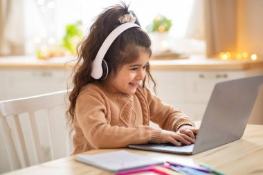 Cute Little Preschooler Girl In Wireless Headphones Using Laptop At Home clipart