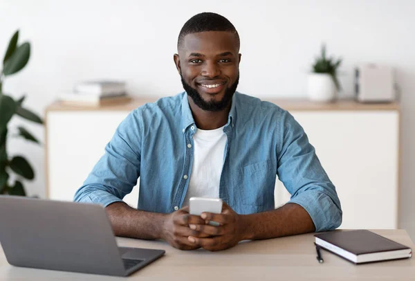 Uśmiechnięty czarny przedsiębiorca siedzi przy biurku w biurze, trzymając smartfon — Zdjęcie stockowe