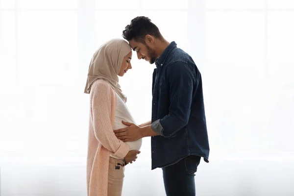 Joven pareja musulmana religiosa esperando un bebé, de pie juntos cerca de la ventana en casa — Foto de Stock