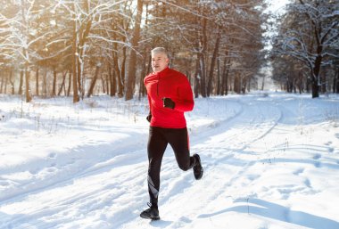 Athletic mature man in sportswear jogging at snowy winter park. Healthy senior runner sprinting outdoors in cold weather clipart