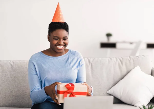 Χαμογελώντας μαύρη γυναίκα στο καπέλο κρατώντας κουτί δώρου χρησιμοποιώντας το laptop — Φωτογραφία Αρχείου