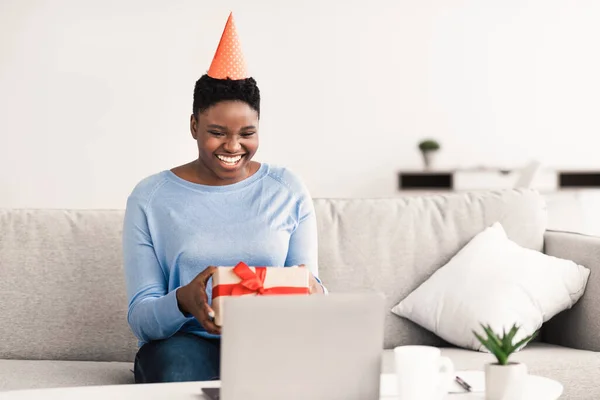Emocionado mujer negra en sombrero sosteniendo caja de regalo usando el ordenador portátil — Foto de Stock