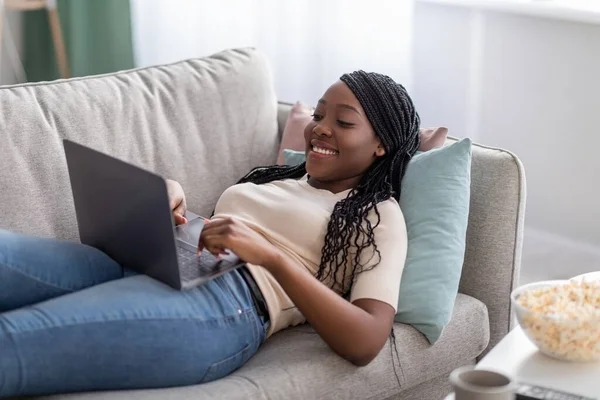 Glädjande afrikansk dam ligger på soffan hemma, med hjälp av laptop — Stockfoto