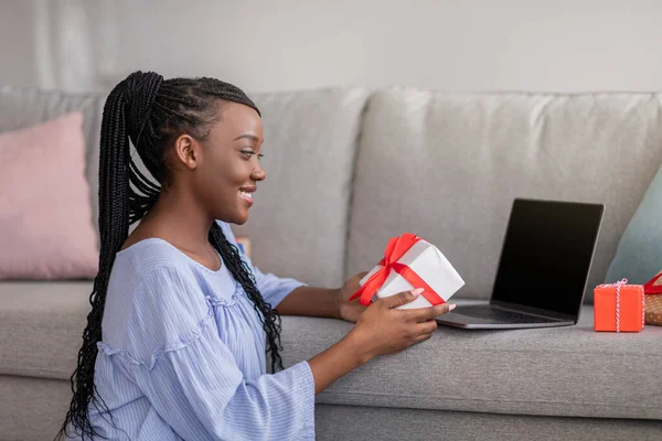 Smiling black woman holding gift, using laptop with blank screen