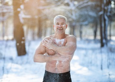 Athletic mature guy tempering his body with snow, developing resistance to cold at white winter forest clipart