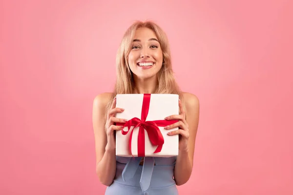 Allegra signora millenaria in grazioso vestito che mostra scatola avvolta con regalo per il giorno della donna o compleanno su sfondo rosa — Foto Stock